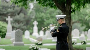 A person standing by a gravestone, holding a bouquet of flowers and a small bag of supplies.